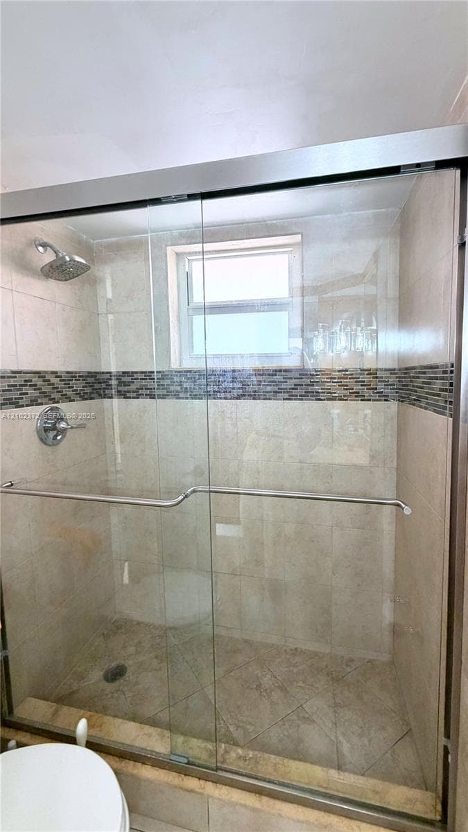 Walk-in shower featuring a sliding glass door, neutral tile surround, a decorative stone mosaic accent band, a built-in window, and a stainless steel shower head with integrated controls
