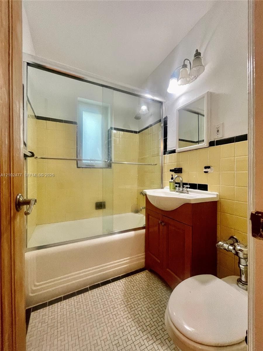 Bathroom featuring a vanity with an integrated basin, a soaking tub with sliding glass doors, tiled walls with decorative trim, and geometric patterned floor tile