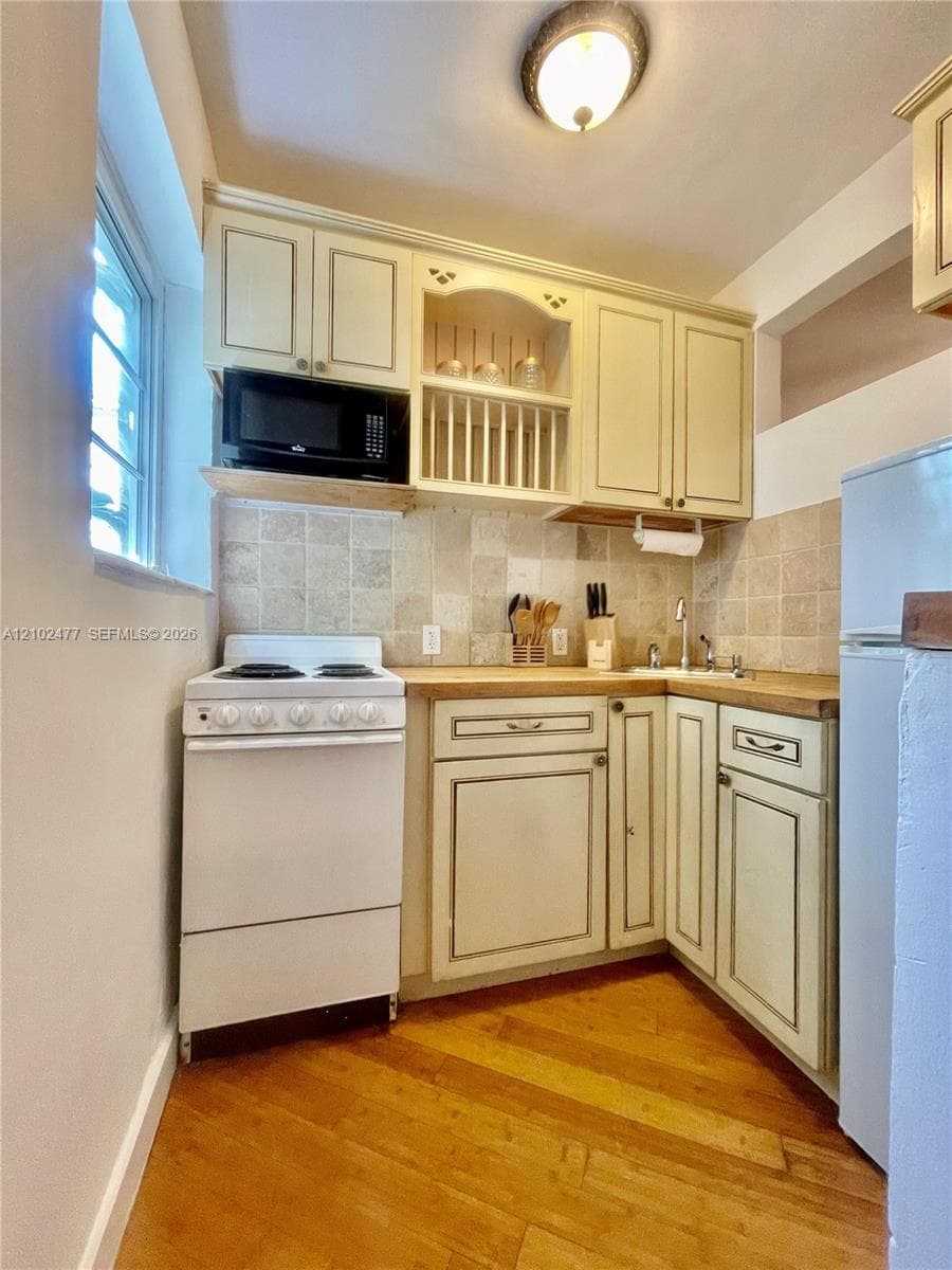 Kitchenette featuring off-white shaker-style cabinetry with open plate rack, stone tile backsplash, wood-finish countertop, and wood flooring