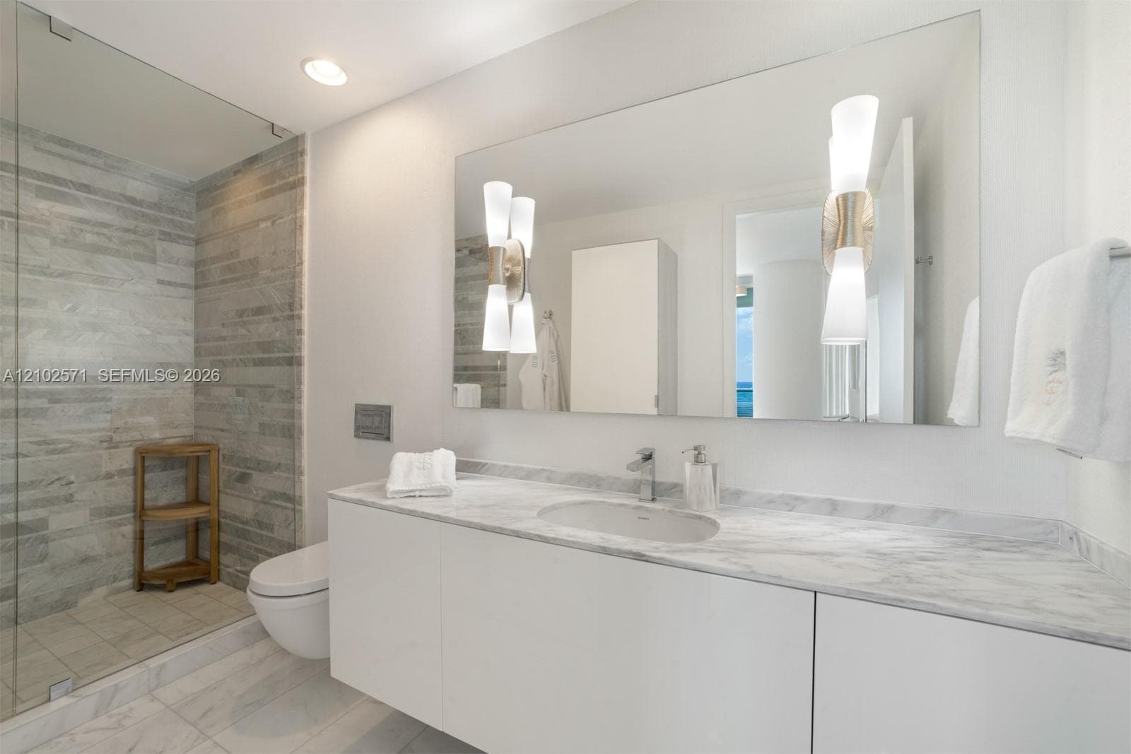 Floating vanity with integrated sink, stone-surface countertop, walk-in glass-enclosed shower featuring horizontal stone-tiled walls, wall-mounted toilet, and dual decorative light fixtures