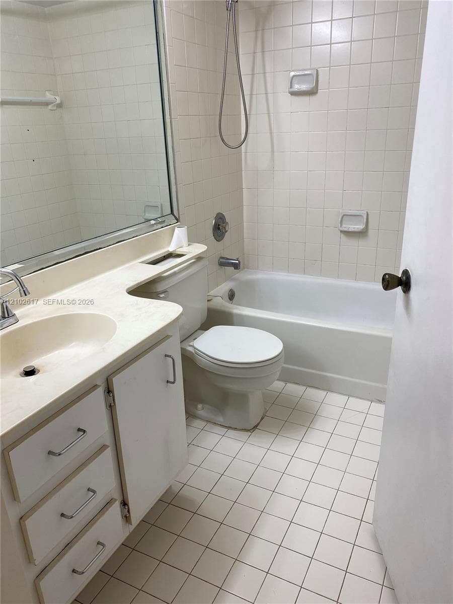 Bathroom featuring white ceramic wall tiles, a built-in bathtub with a shower head, a white vanity with integrated sink, and square floor tiles
