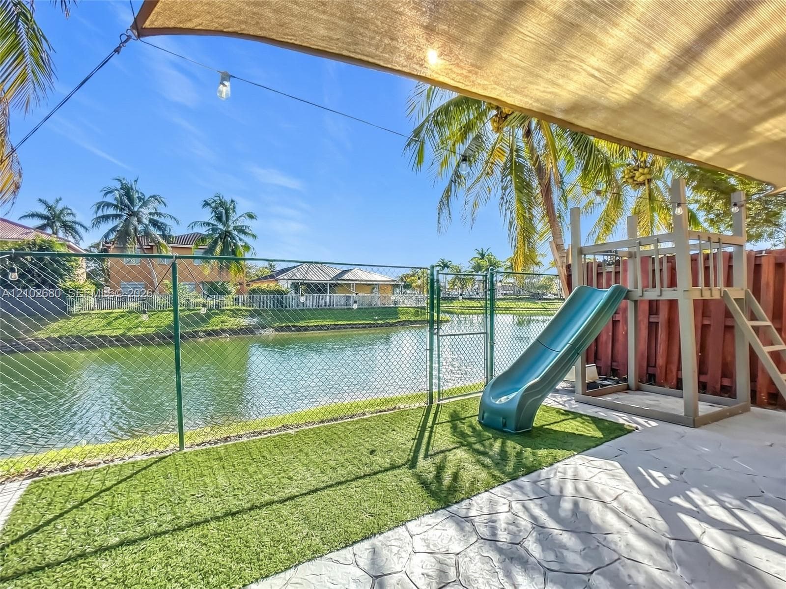 Waterfront backyard featuring a stamped concrete patio, synthetic turf, metal chain-link fencing, and a permanent play structure under a shade sail