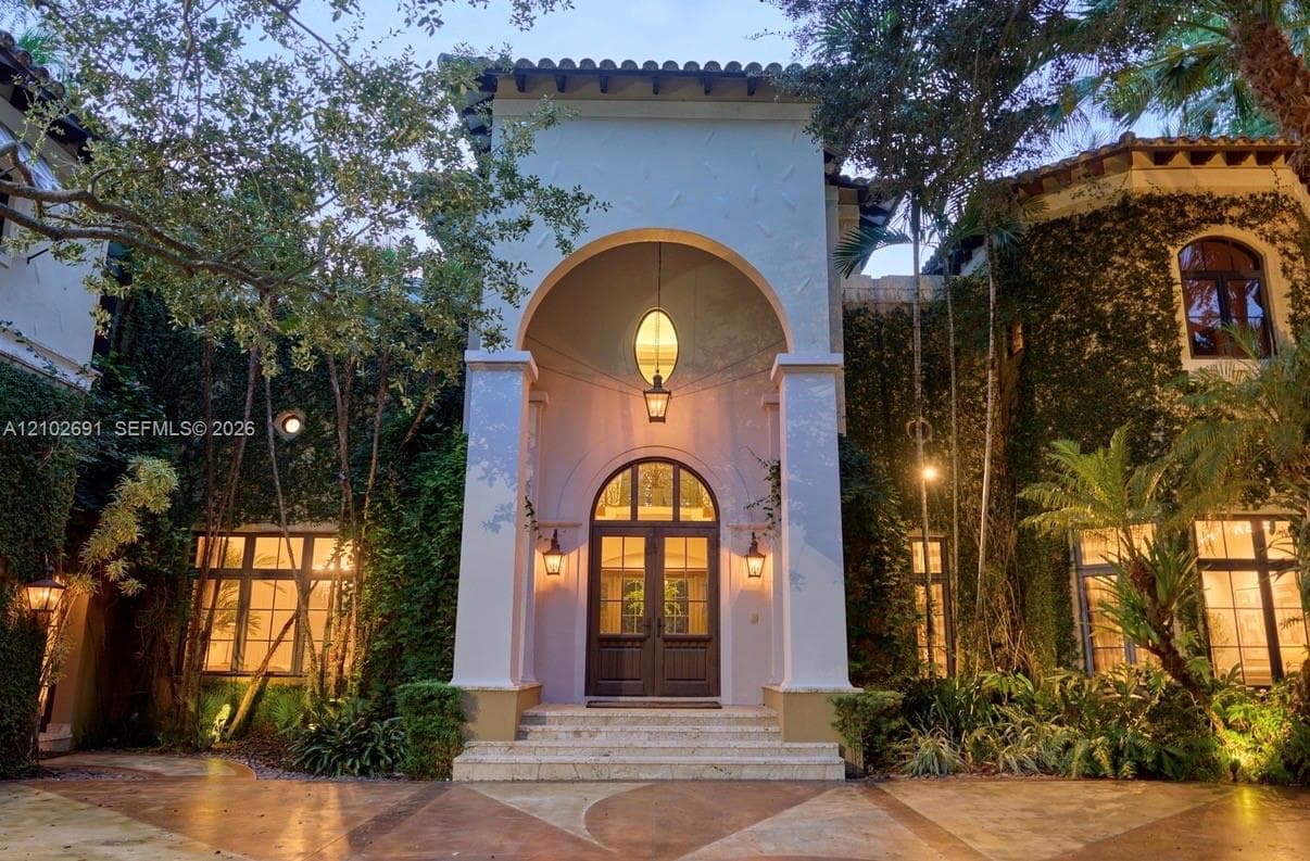 Mediterranean-inspired exterior featuring a grand arched entryway, stucco facade, decorative iron-caged pendant lighting, and stone steps leading to double entry doors