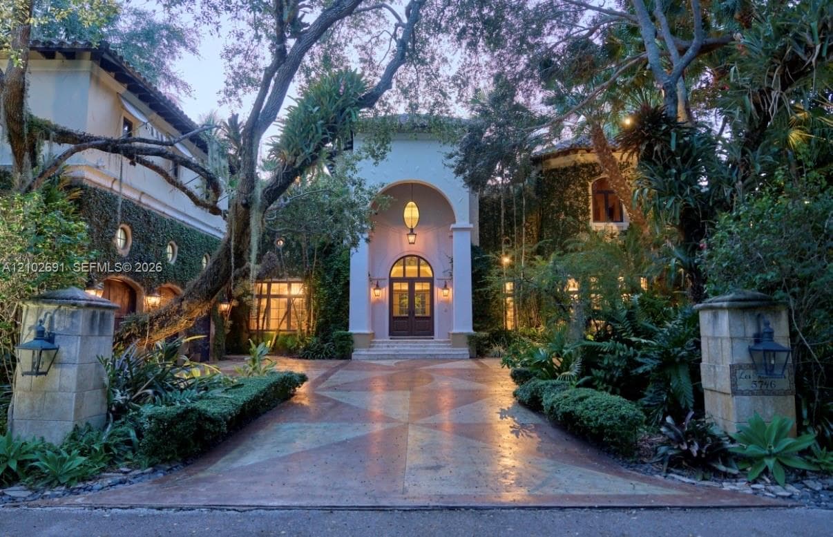 Stucco exterior residence featuring an arched portico entrance, geometric patterned driveway, stone entry pillars with mounted lanterns, and exterior climbing vegetation