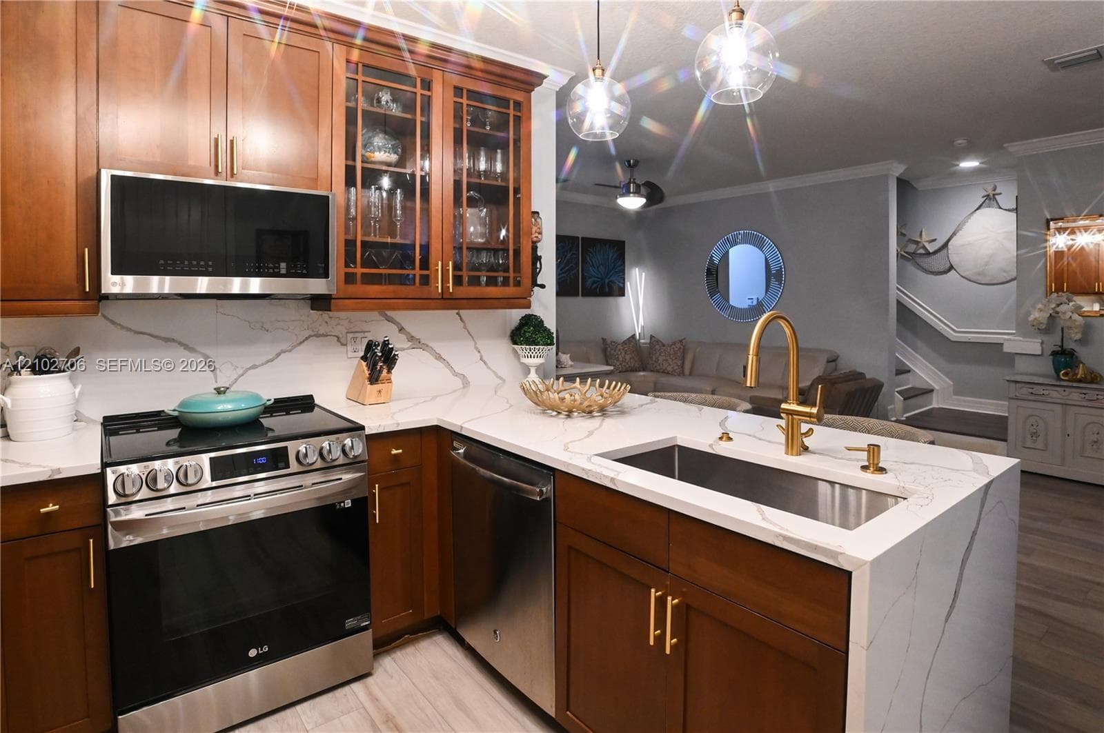 Kitchen featuring warm-toned cabinetry, a waterfall-edge countertop with integrated backsplash, a stainless steel slide-in range, a single-basin undermount sink with a brass-finish faucet, and glass-front upper cabinet displays