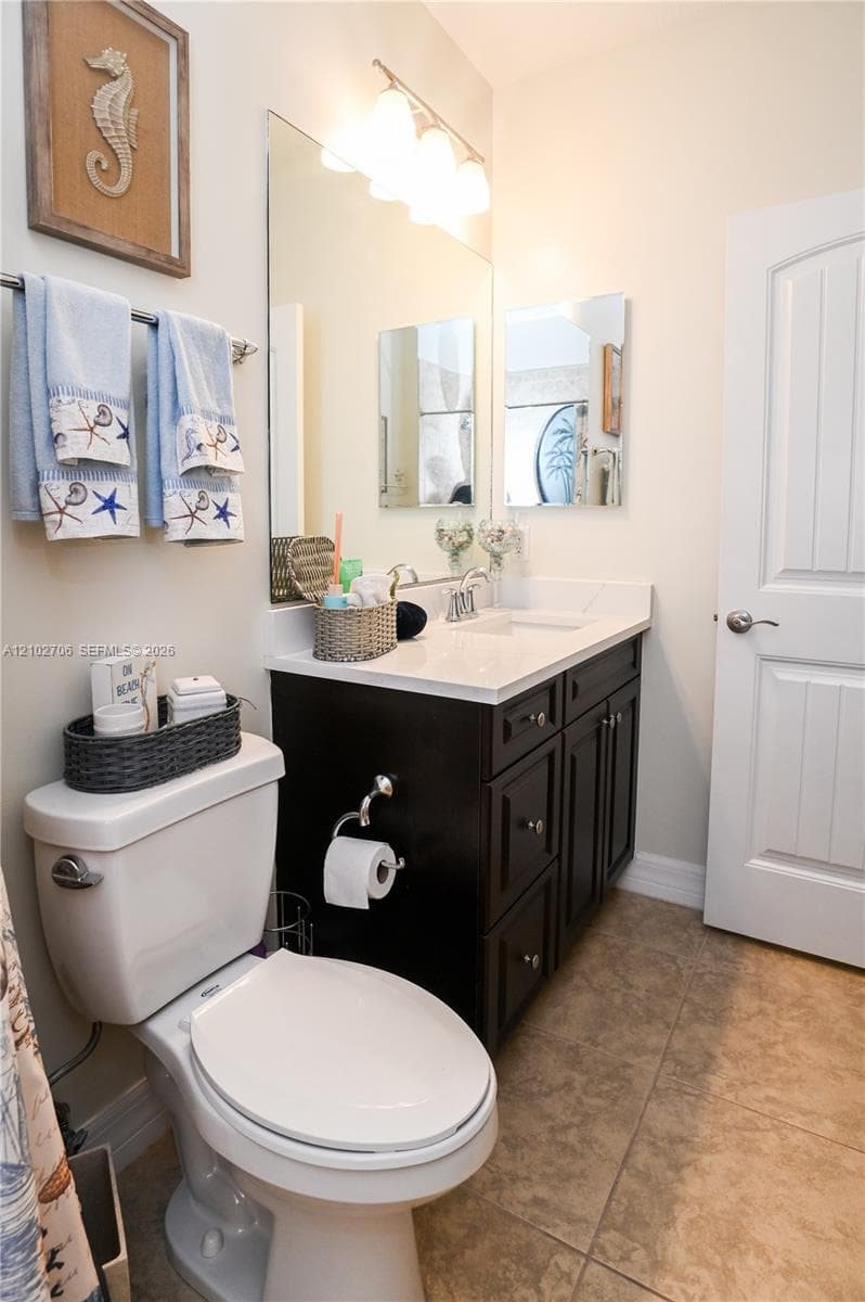 Bathroom vanity with dark cabinetry, stone-finish countertop, integrated sink, and wall-mounted vanity lighting, complemented by tile flooring