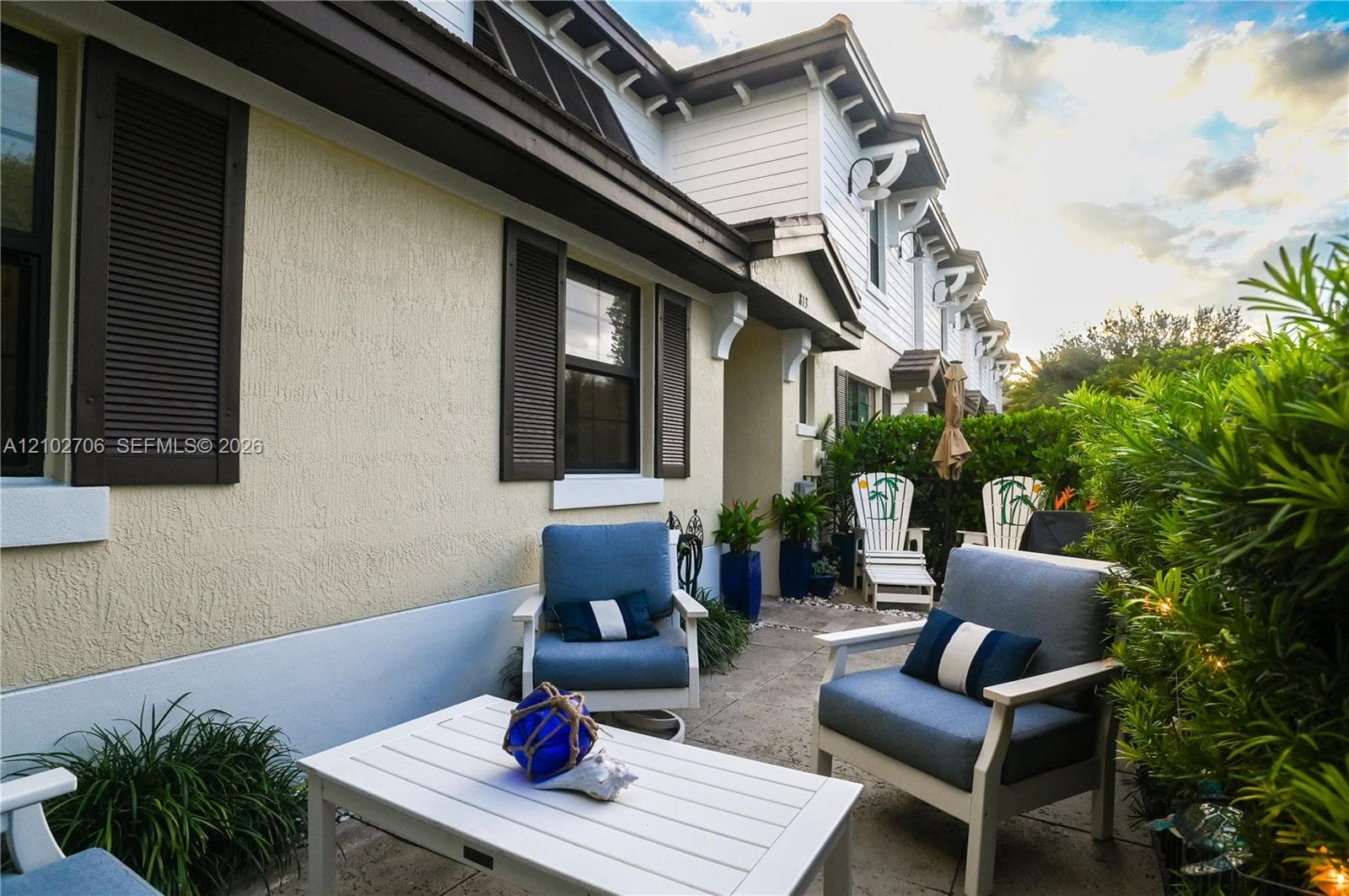 Exterior facade featuring light-colored stucco siding, dark-toned shutters, architectural roof detailing, and a stone-paved patio area bordered by mature shrubbery