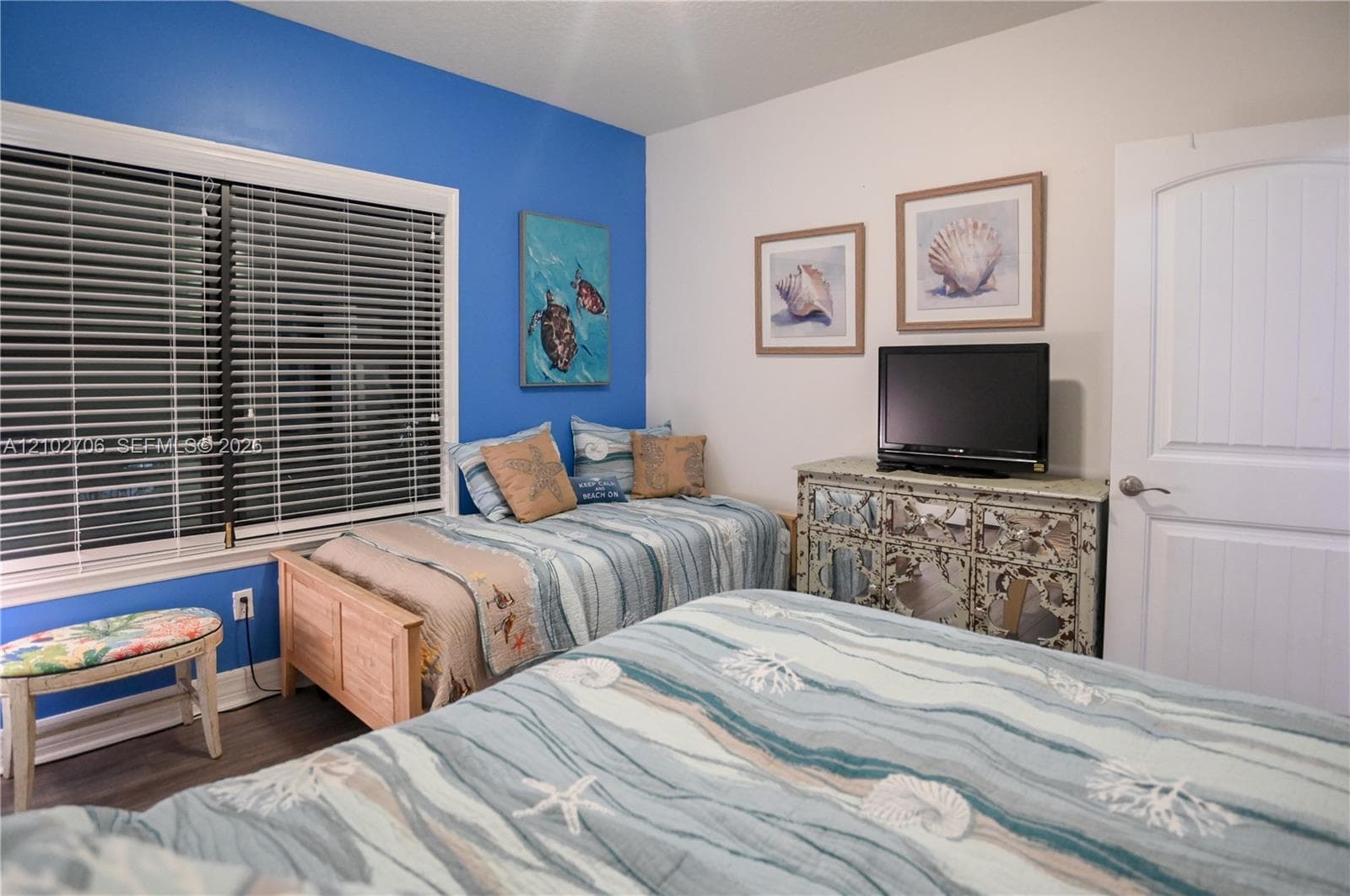 Bedroom featuring wood-finish flooring, a vibrant blue accent wall, horizontal window blinds, and a paneled interior door with a brushed metal handle