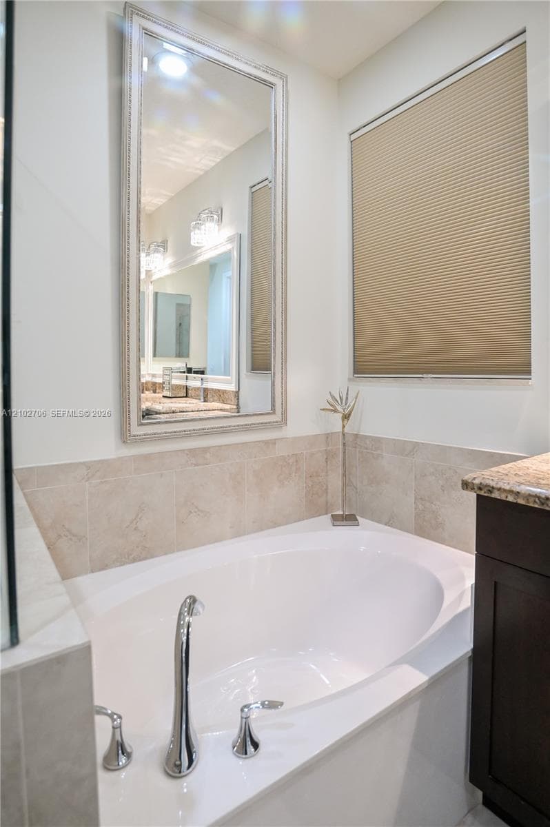 Soaking tub featuring chrome fixtures, tile surround, framed wall-mounted mirror, cellular shade, and dark-finish cabinetry with a stone-top vanity