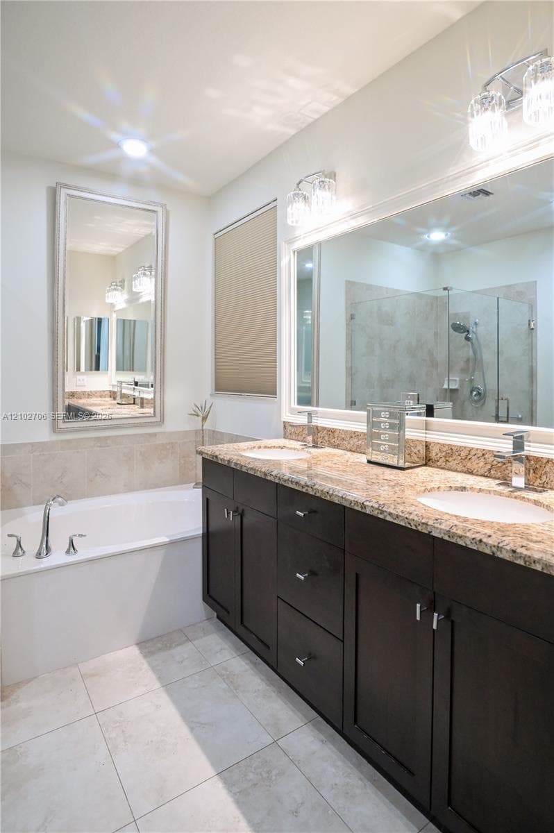 Double vanity with stone-surface countertop, under-mount sinks, dark-finish cabinetry, drop-in soaking tub, and walk-in shower with glass enclosure