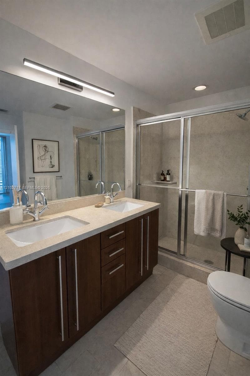 Bathroom featuring a dual-sink vanity with light-toned stone-finish countertop, dark wood-finish cabinetry, floor-to-ceiling mirror with integrated lighting, and a glass-enclosed shower with tiled surround