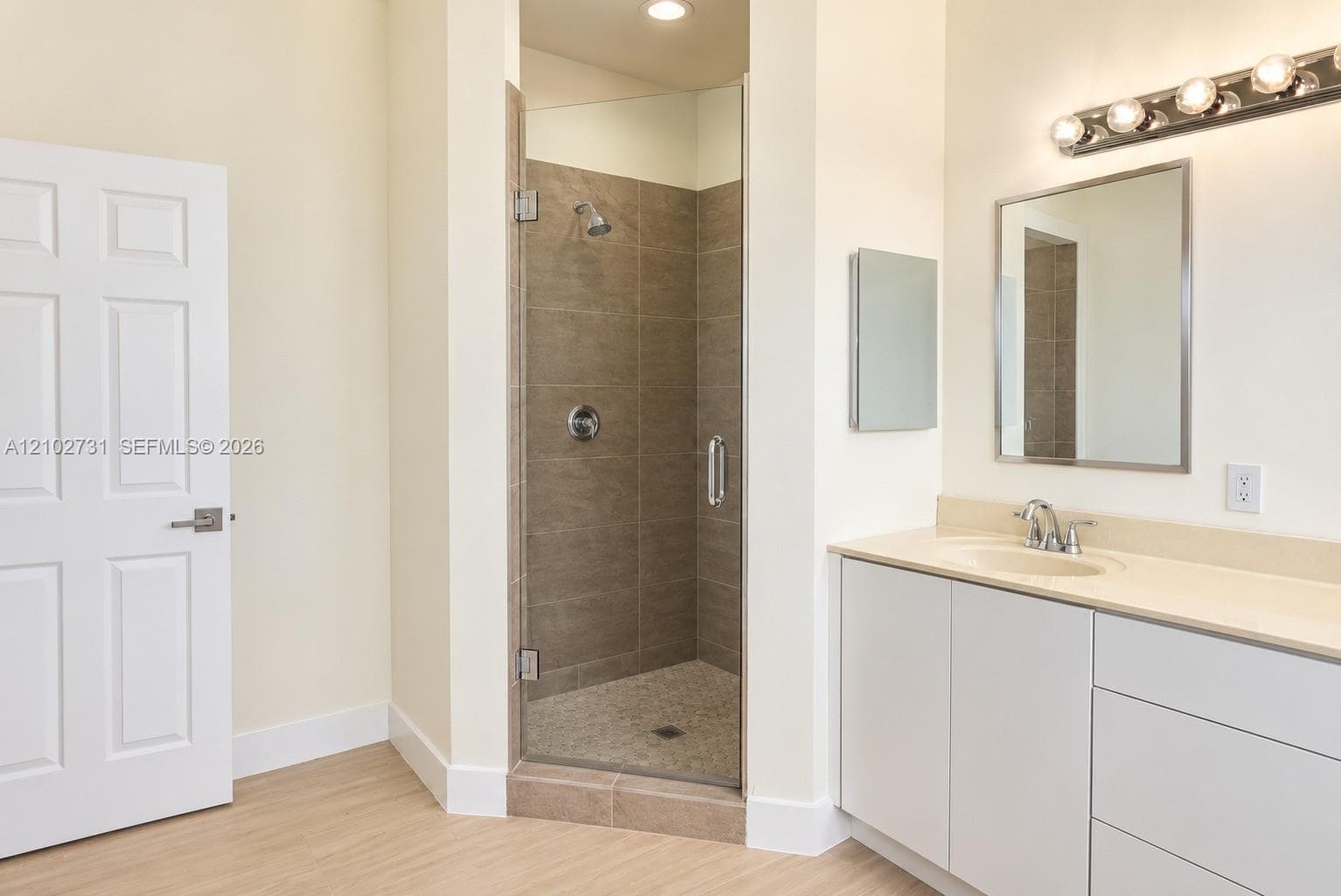 Enclosed shower with full-height tile surround and glass door, integrated vanity with solid surface countertop, rectangular wall-mounted mirror, and multi-bulb polished finish vanity lighting