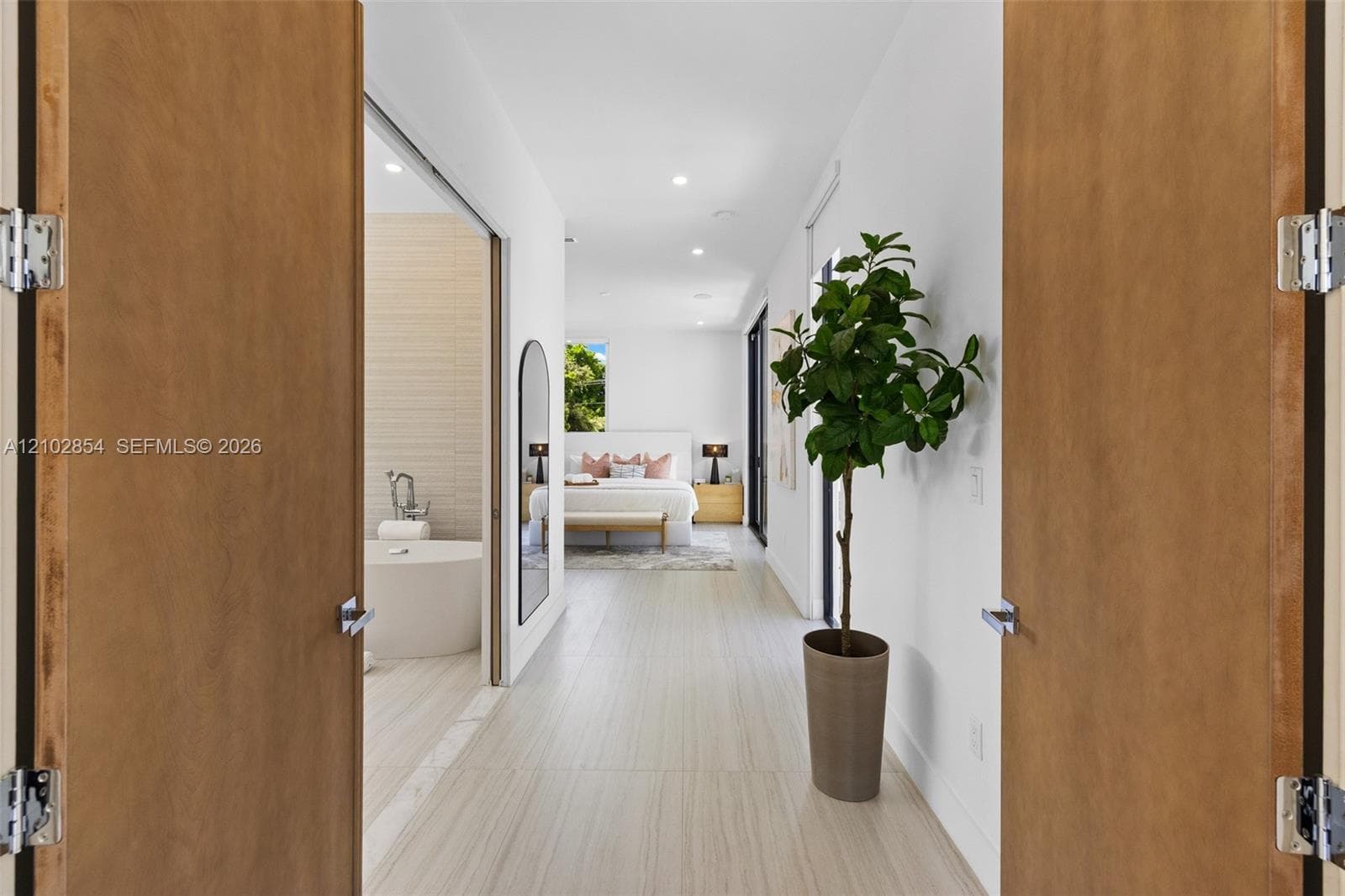 Double wood-finish doors opening to a space with neutral large-format tile flooring, recessed lighting, and a freestanding soaking tub