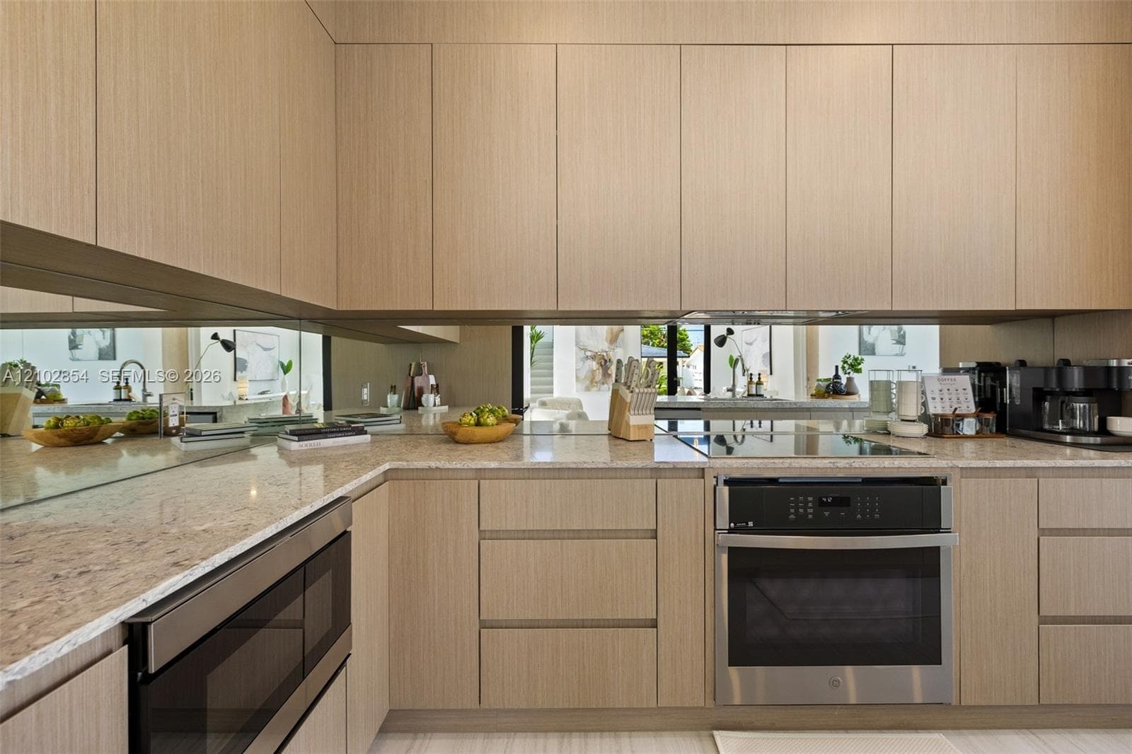Modern kitchen featuring flat-panel wood-finish cabinetry, neutral stone countertops, a mirrored backsplash, built-in stainless steel oven, and a flat-surface cooktop
