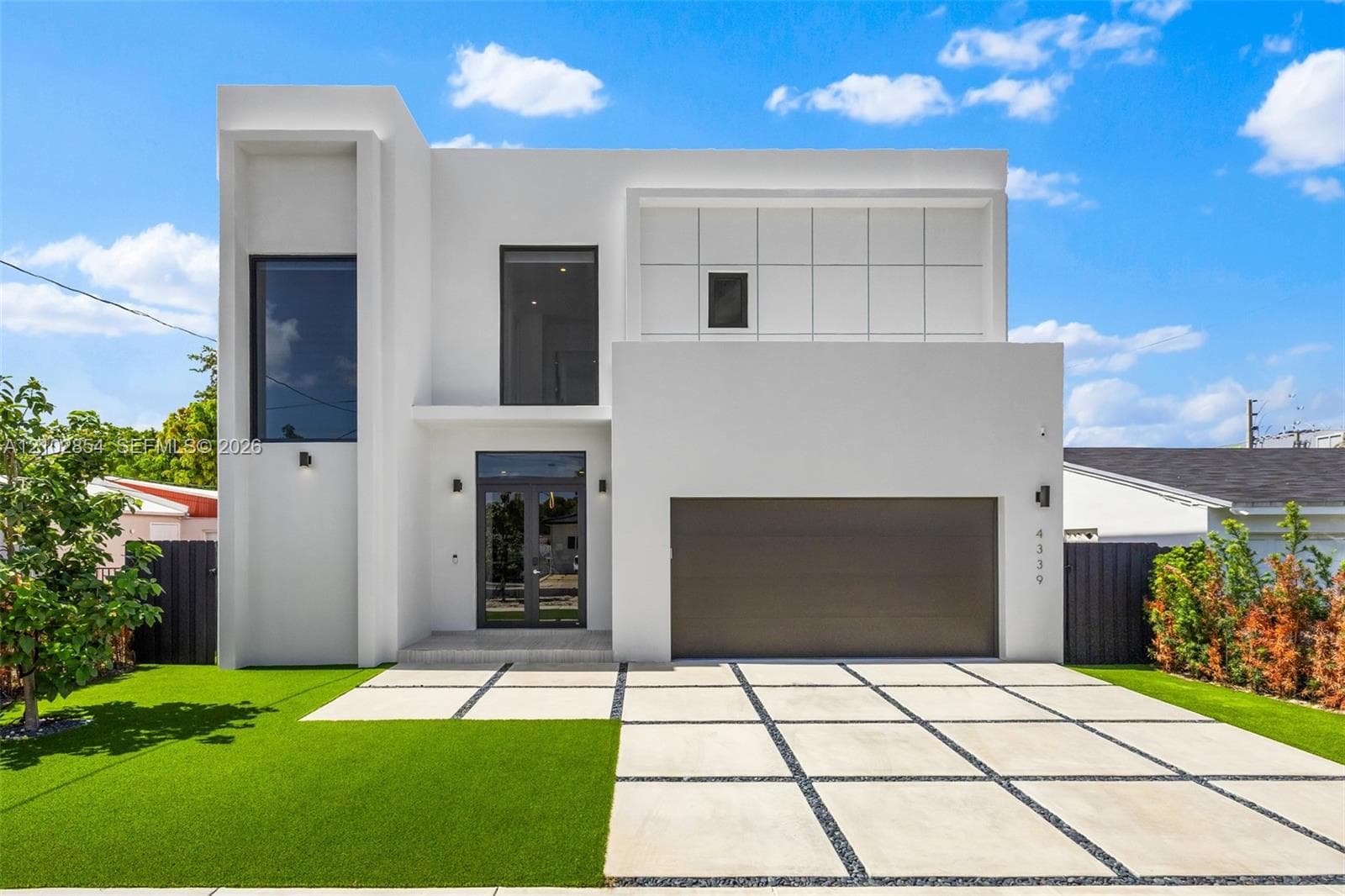 Contemporary architectural exterior featuring a smooth white stucco facade, geometric grid-patterned concrete driveway, inset glass-panel front entry door, large-format vertical windows, and square-tiled upper-level cladding