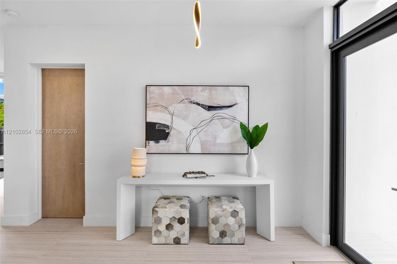 Minimalist interior featuring light wood-finish flooring, a solid core wood-finish door, a sculptural contemporary pendant light fixture, and a large-format glass door with a matte black frame