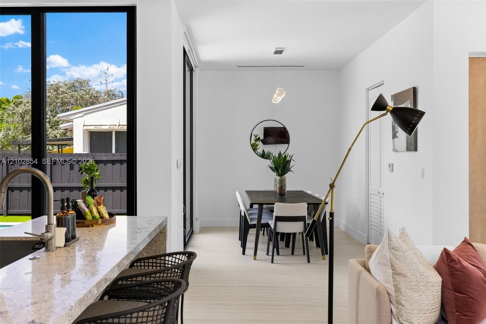 Open-concept interior featuring light-toned flooring, expansive floor-to-ceiling black-framed windows, and a kitchen island with a stone-finish countertop and undermount sink