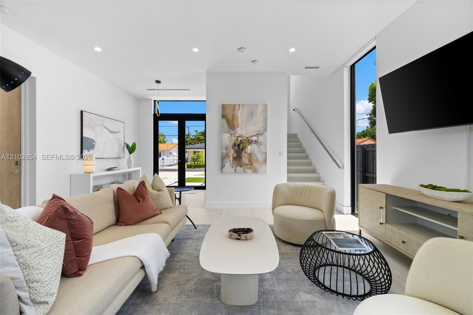 Open-concept living space featuring high ceilings, recessed lighting, full-height windows, a glass-paned entry door, and a floating staircase with a metal handrail