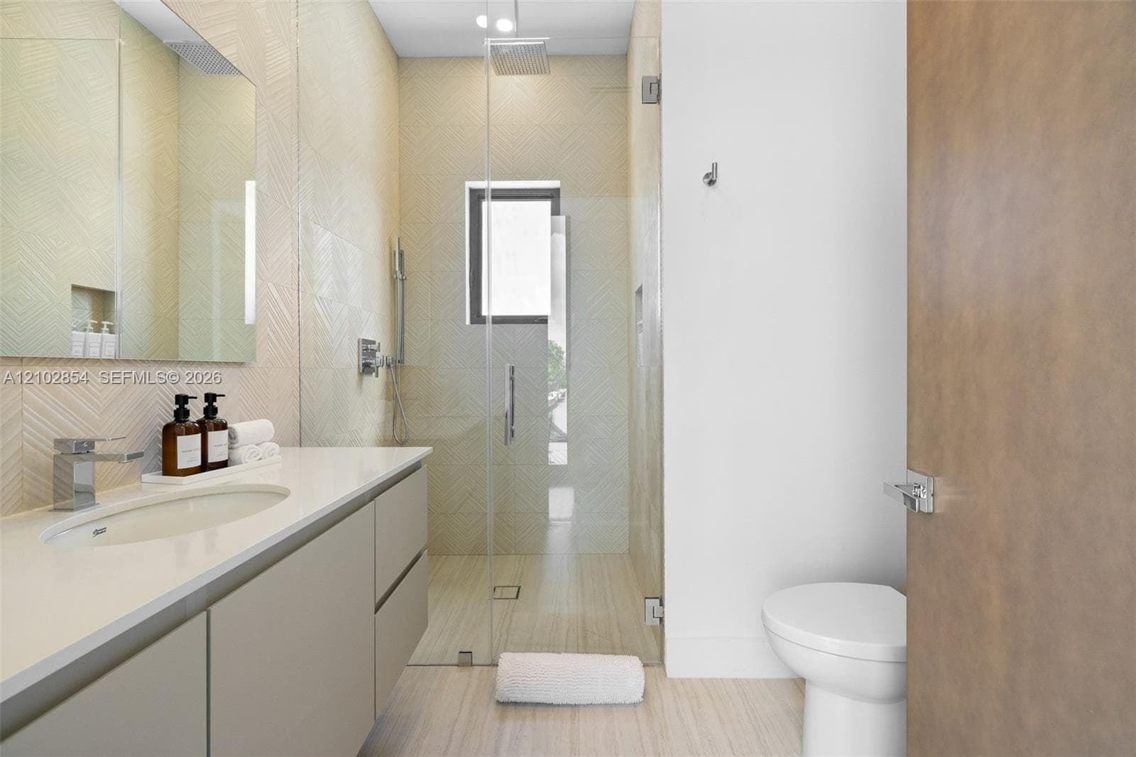 Floating vanity with a smooth-surface countertop, integrated rectangular sink, wall-mounted mirror, glass-enclosed walk-in shower with textured tile surround, and neutral large-format floor tiling