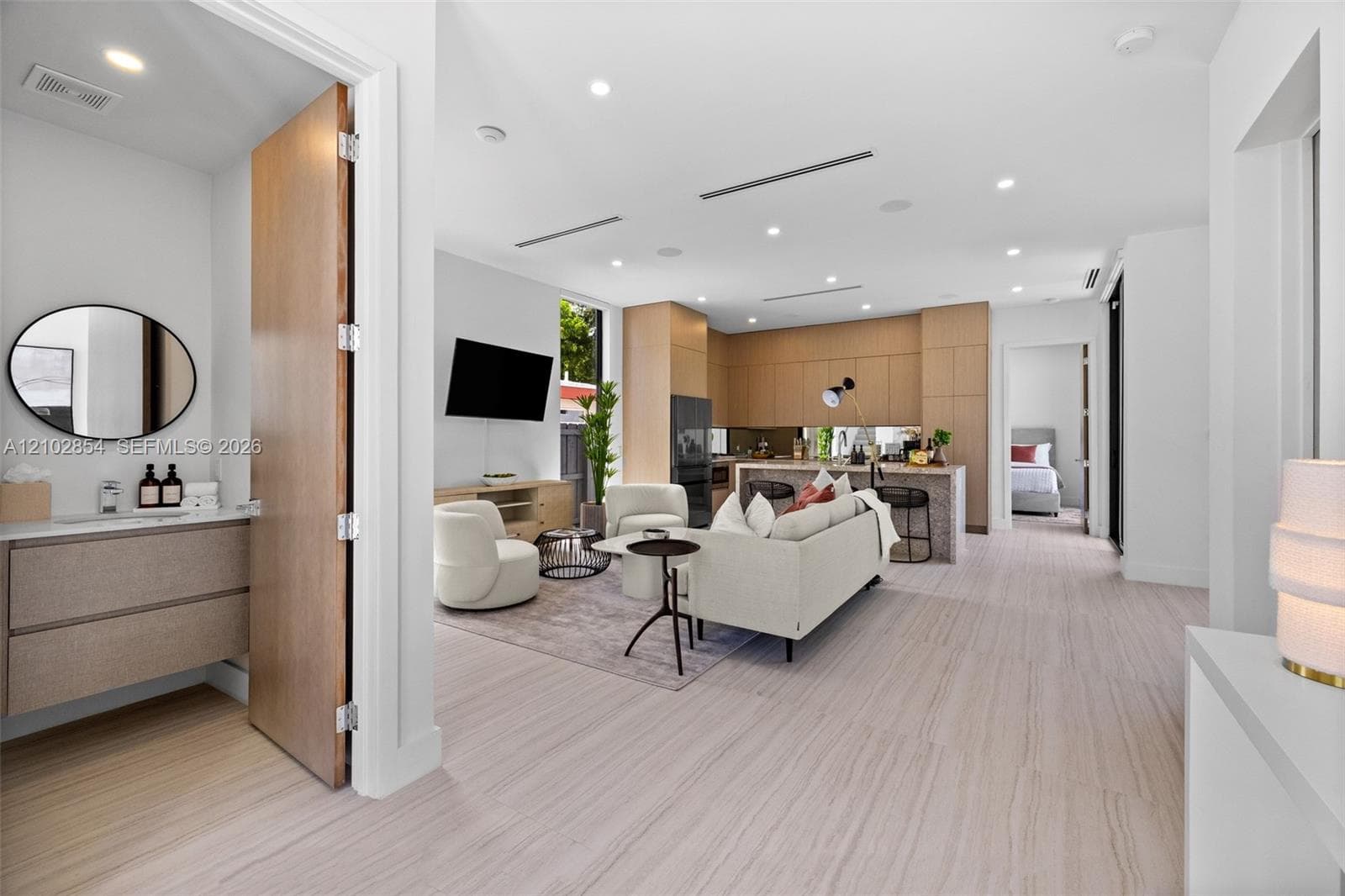 Open-concept interior featuring light wood-finish flooring, recessed lighting, kitchen cabinetry with flat-panel wood-finish doors, a center island with stone-finish waterfall countertop, and a vanity with a circular mirror