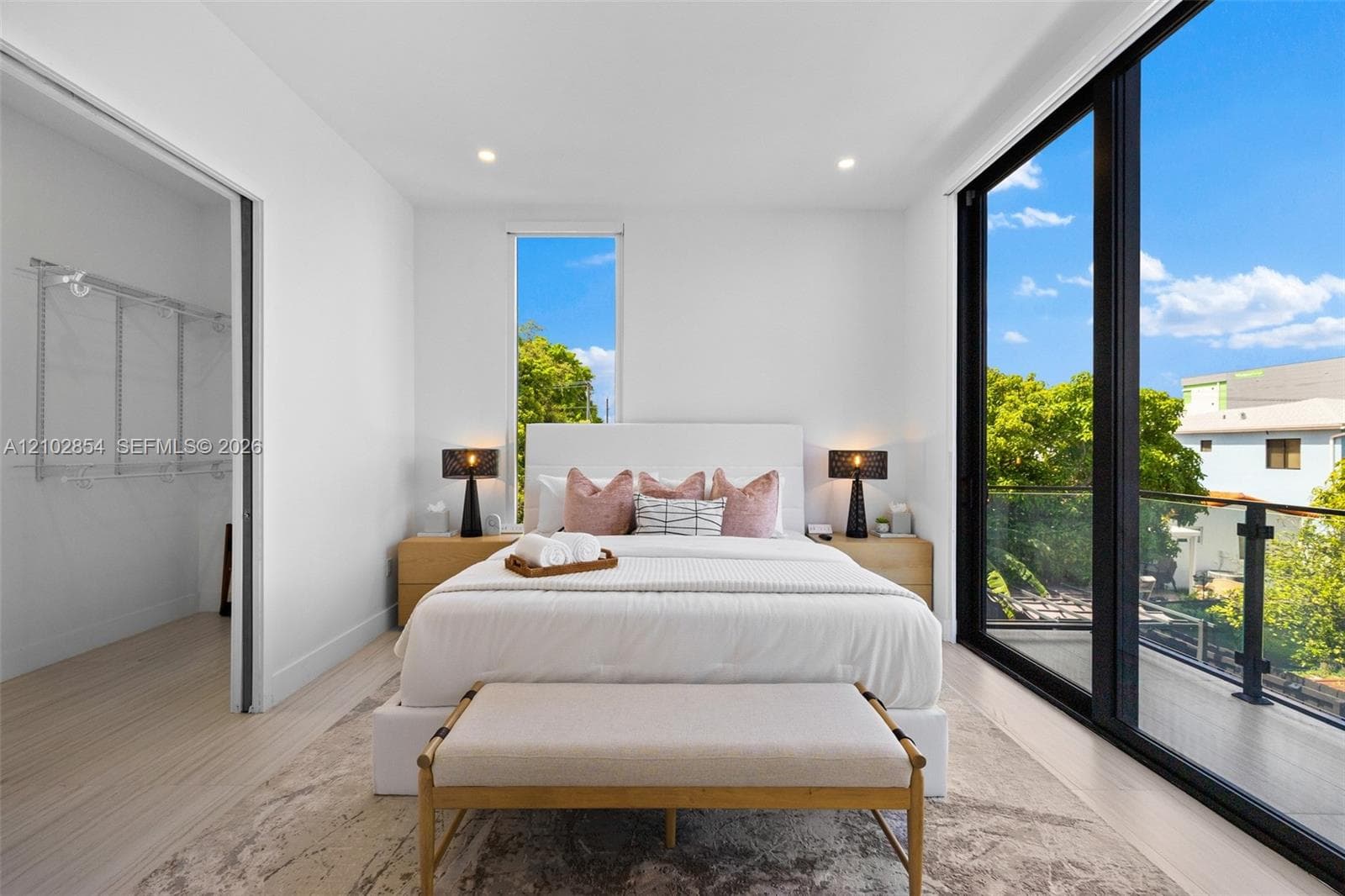 Full-height glass sliding doors, private balcony with metal railing, light-tone wood-finish flooring, recessed ceiling lighting, and integrated walk-in closet shelving