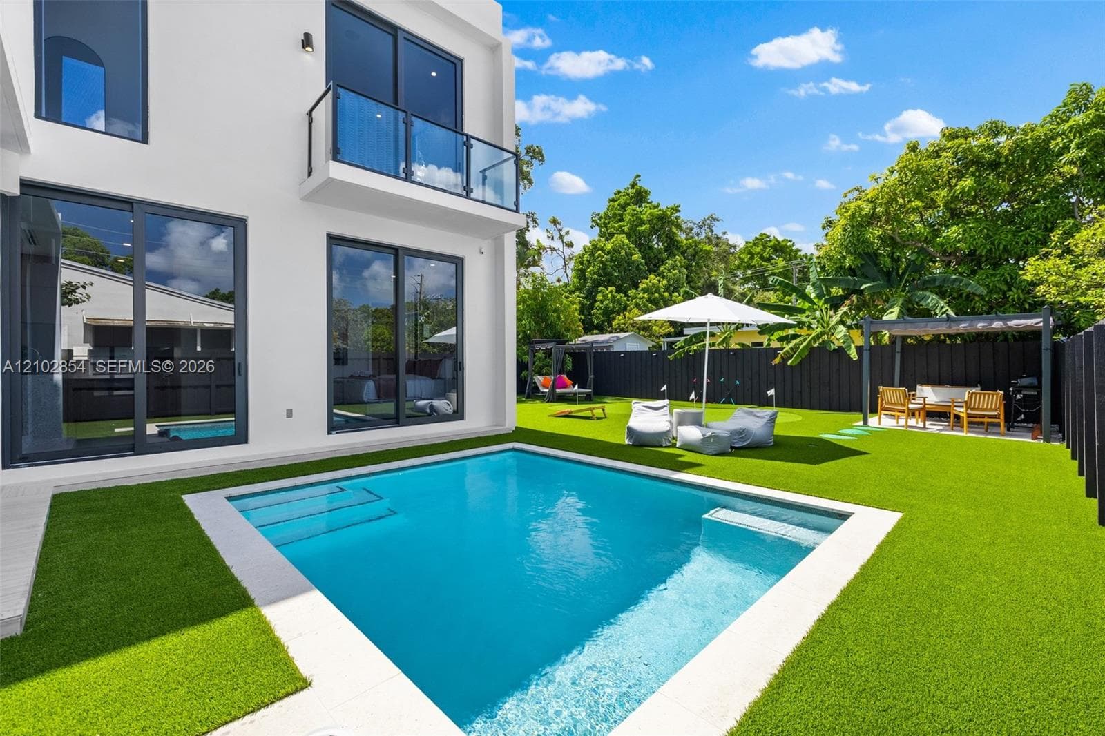 Modern exterior featuring a geometric in-ground pool, synthetic turf lawn, second-story glass-railed balcony, and black sliding glass doors