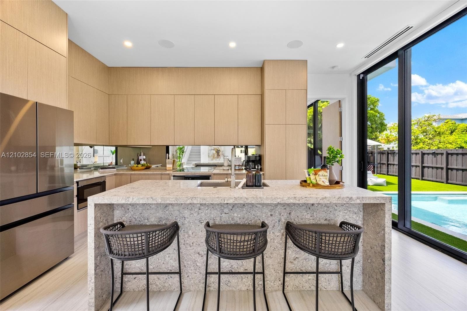 Flat-panel cabinetry, waterfall-edge stone island with integrated sink, floor-to-ceiling glass sliding doors, stainless steel refrigerator, and recessed lighting