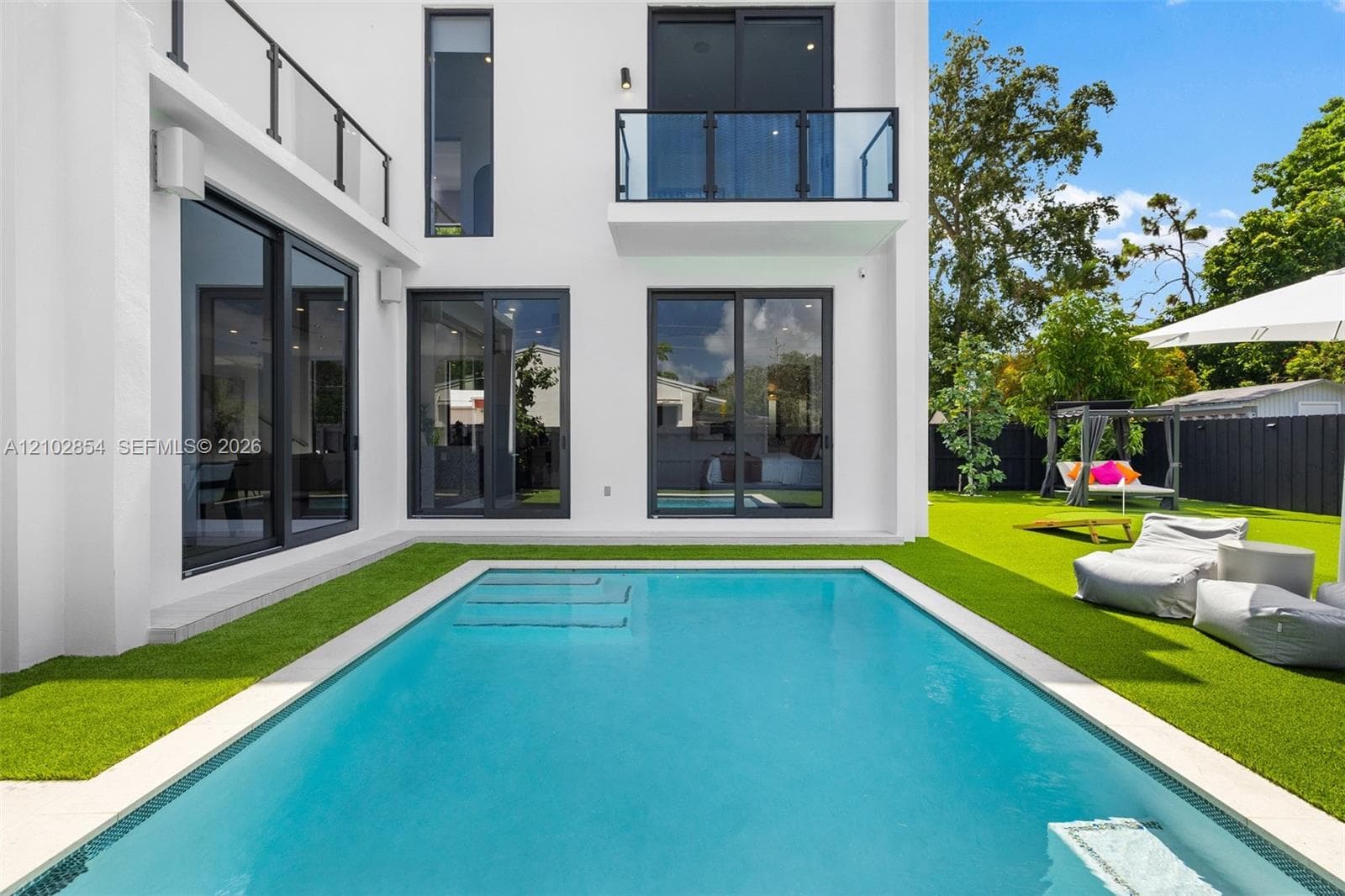 Modern exterior featuring floor-to-ceiling glass sliding doors, upper-level balconies with glass railings, a swimming pool with integrated stairs, and artificial turf hardscaping