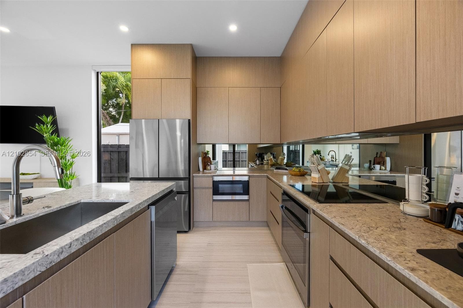 Contemporary kitchen featuring full-height flat-panel cabinetry, light stone-finish countertops, stainless steel appliances, mirror backsplash, and recessed lighting
