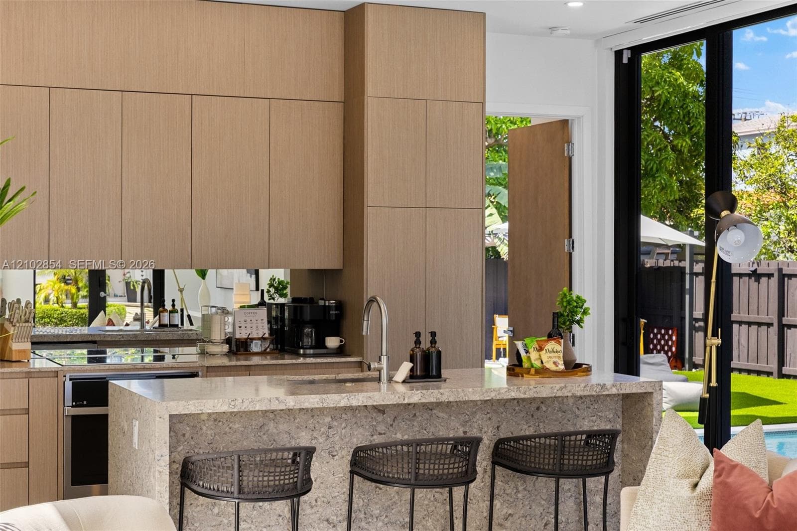Kitchen featuring flat-panel wood-finish cabinetry, a waterfall-edge stone island with a stainless steel faucet, a mirrored backsplash, and floor-to-ceiling glass doors