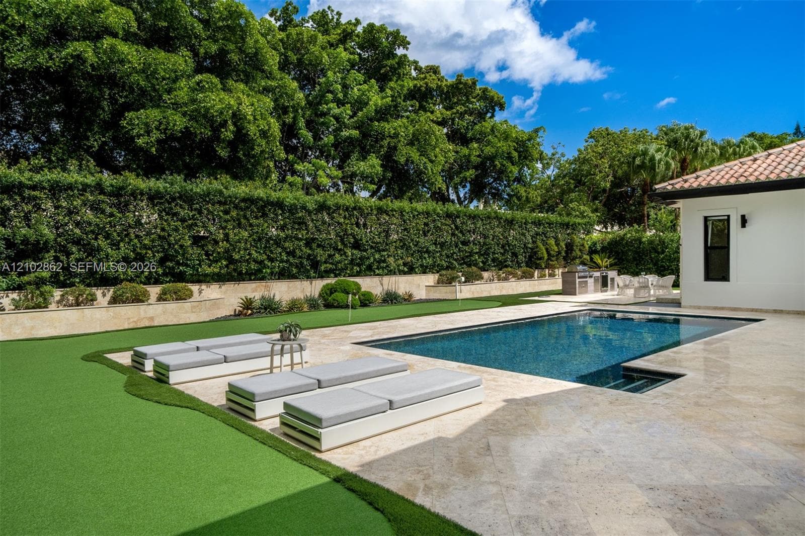 Outdoor living area featuring a custom swimming pool, travertine paver patio, synthetic turf lawn, built-in outdoor kitchen, and perimeter landscaping