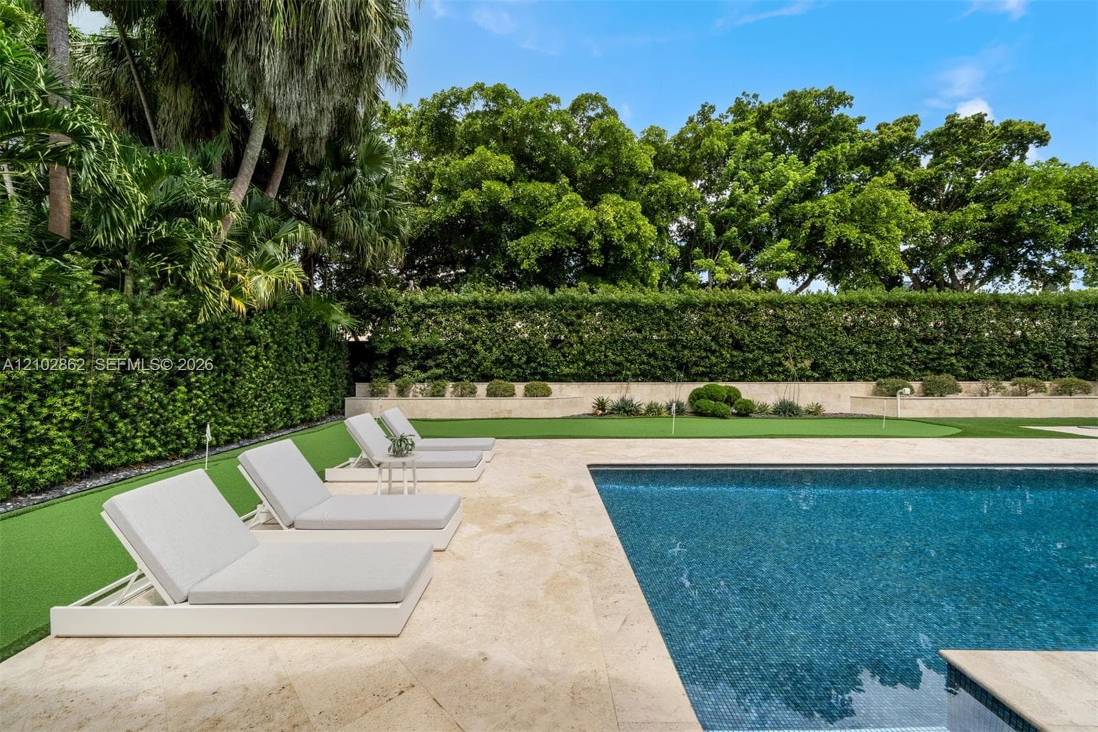 Private outdoor space featuring a rectangular swimming pool with stone-tiled decking, a manicured artificial turf putting green, and perimeter hedges