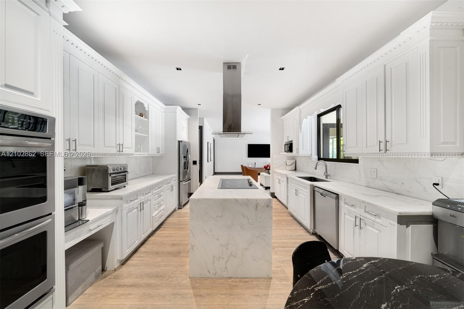 Gourmet kitchen featuring white shaker cabinetry, a waterfall-edge center island with integrated cooktop, stone-finish countertops, stainless steel wall-mounted oven, and wood-finish flooring