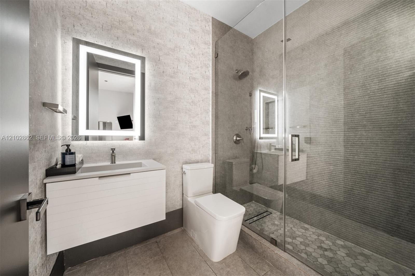 Floating vanity with a smooth-finish countertop, integrated rectangular sink, wall-mounted illuminated mirror, frameless glass-enclosed shower featuring textured tile work, and hexagonal floor tile