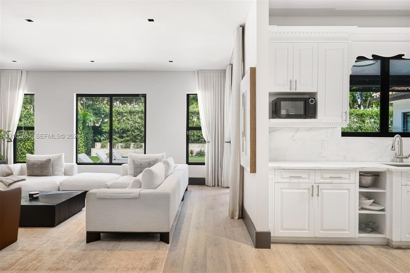 Open-concept interior featuring light wood-finish flooring, white shaker-style cabinetry, solid-surface countertops with stone-pattern veining, and multiple windows with black framing
