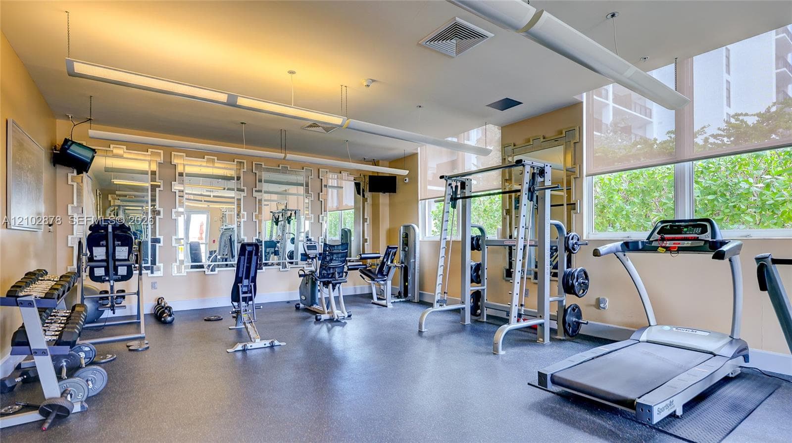 Fitness area featuring rubberized flooring, large wall mirrors with decorative frames, expansive windows with integrated roller shades, and linear pendant lighting
