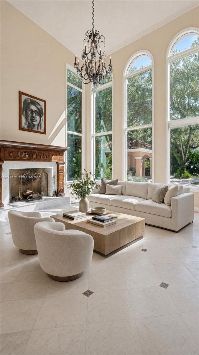 High ceilings featuring an ornate chandelier, a fireplace with a carved wood surround, arched windows, and light-toned tile flooring with dark accent inlays