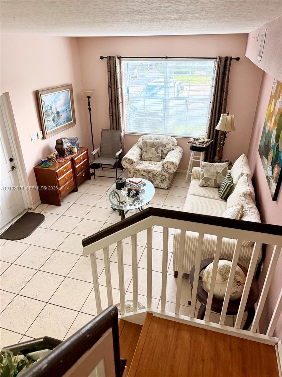 Ceramic tile flooring throughout the main level, natural light from a large window, and staircase featuring a white wooden balustrade with a contrasting handrail