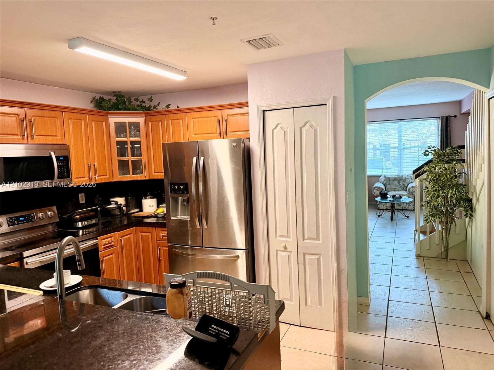 Kitchen featuring wood cabinetry, stainless steel appliances, dark stone-finish countertops, and a double-basin undermount sink, with an arched entryway leading to a tiled area