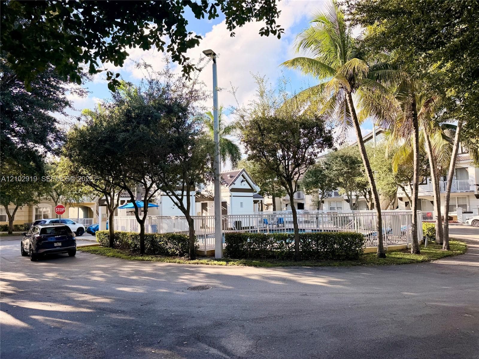 Community swimming area enclosed by white metal fencing, featuring mature palm trees and manicured perimeter shrubbery