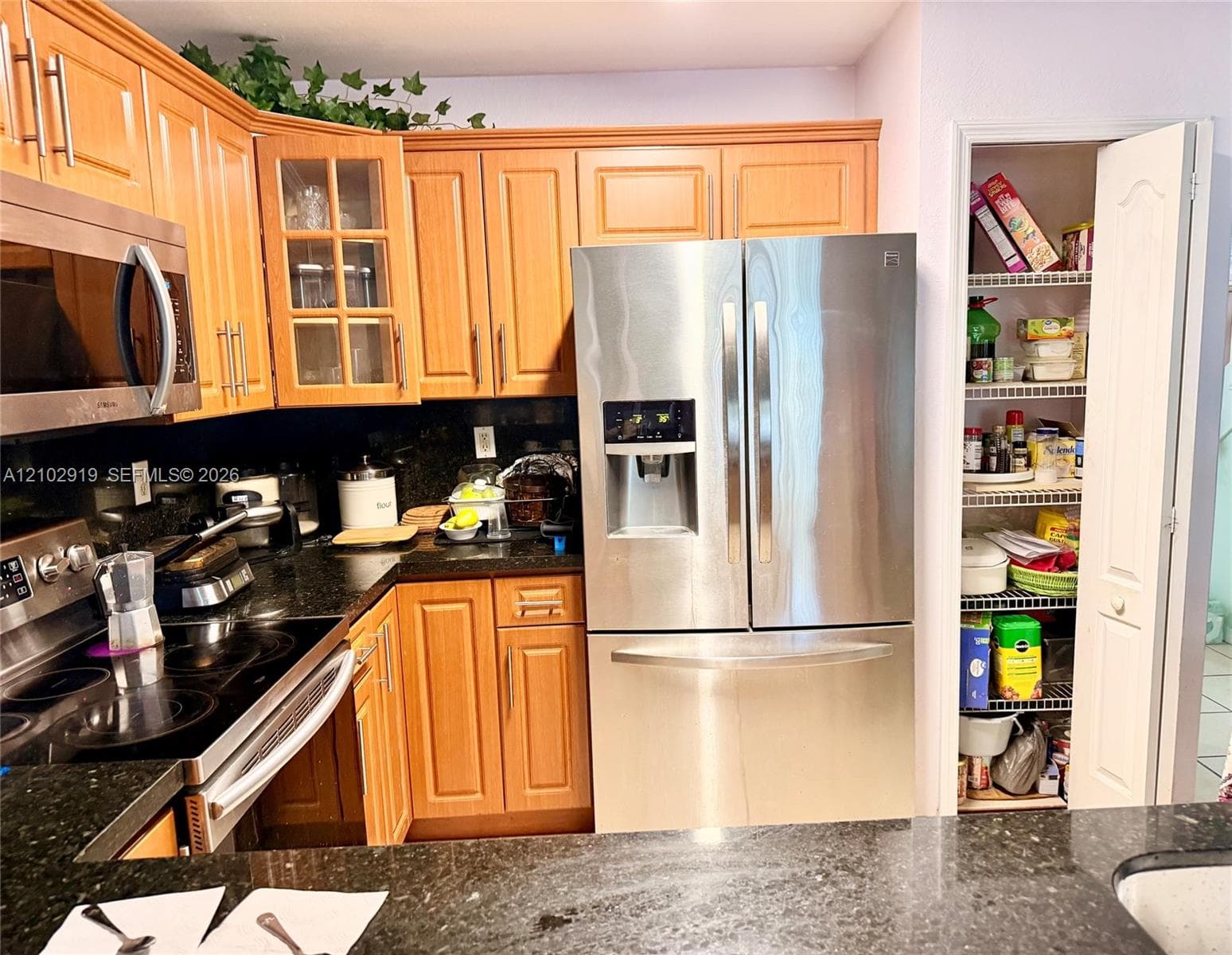 Kitchen featuring wood cabinetry, stone countertops, stainless steel appliances, glass-front upper cabinet doors, and a built-in pantry with wire shelving