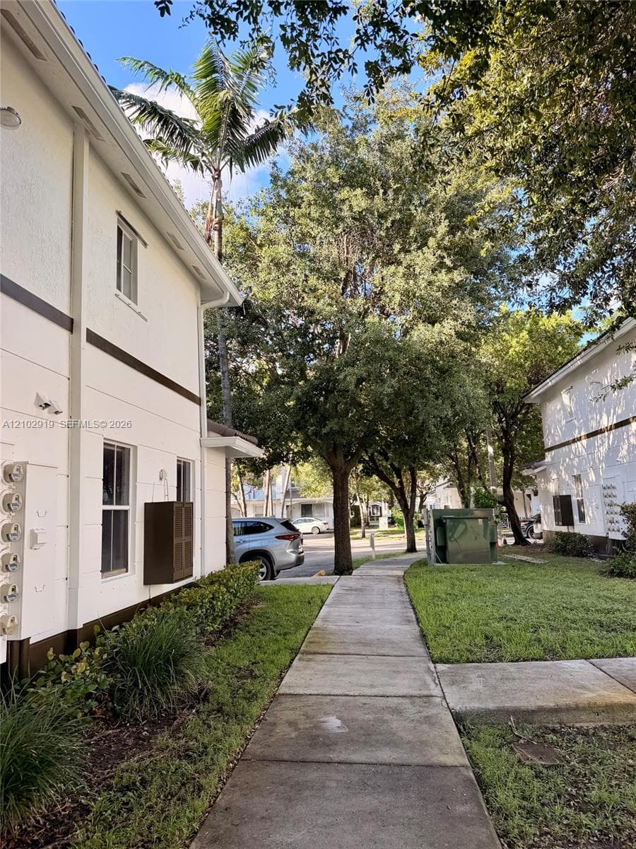 Exterior featuring white stucco siding, concrete sidewalk, mature trees, and landscaped lawn areas