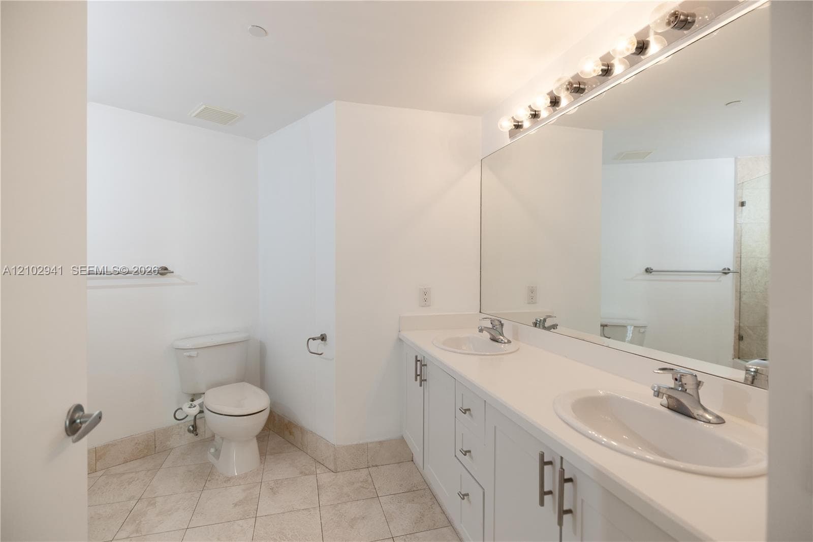 Bathroom featuring a dual-sink vanity with white cabinetry, light-toned countertops, a full-width wall mirror, and neutral-toned tile flooring