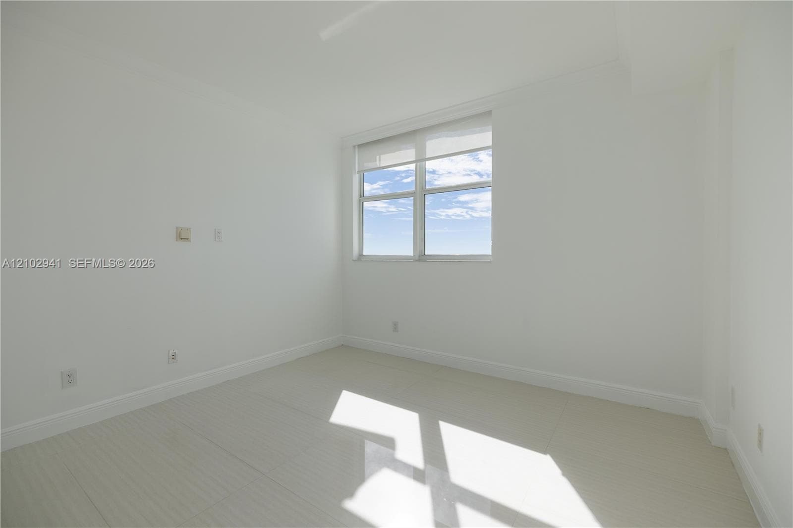 Neutral-toned interior space featuring light-colored tile flooring, smooth wall finishes, and a window with a retractable roller shade