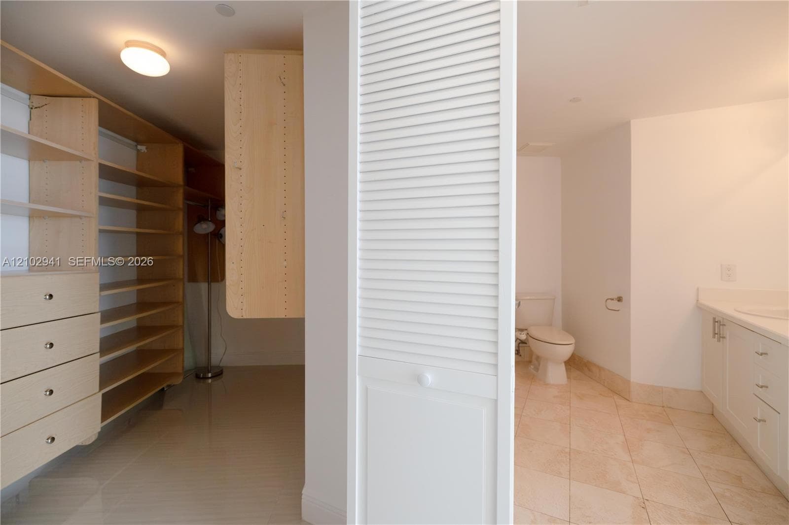 Custom built-in wood shelving and cabinetry with integrated drawers, light-toned tile flooring, white louvered closet doors, and a vanity featuring neutral cabinetry