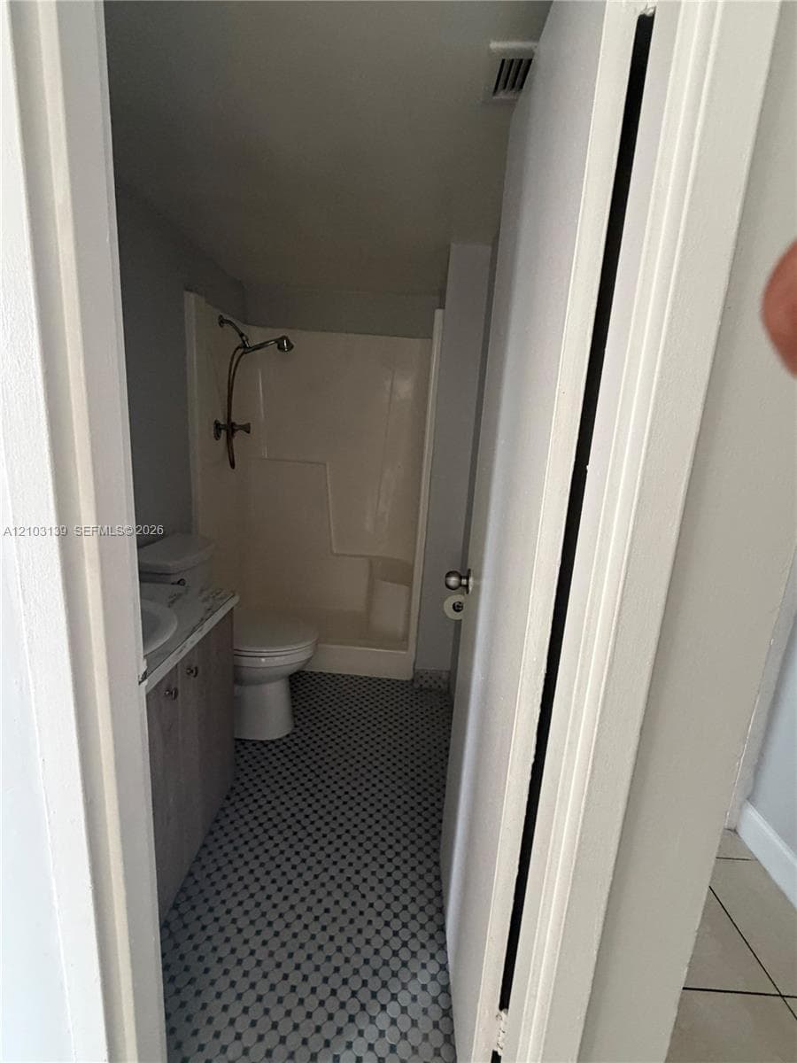 Bathroom featuring patterned tile flooring, a white vanity with dual sinks, a commode, and a shower with a built-in shelf