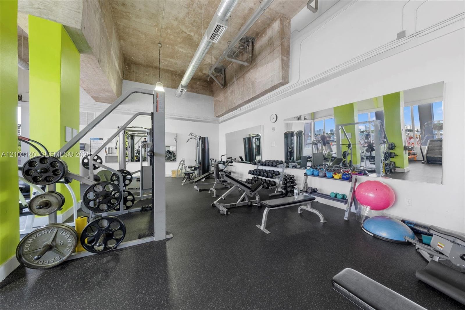 Industrial-style interior featuring exposed concrete ceilings, metallic ductwork, large-scale wall mirrors, and rubberized fitness flooring