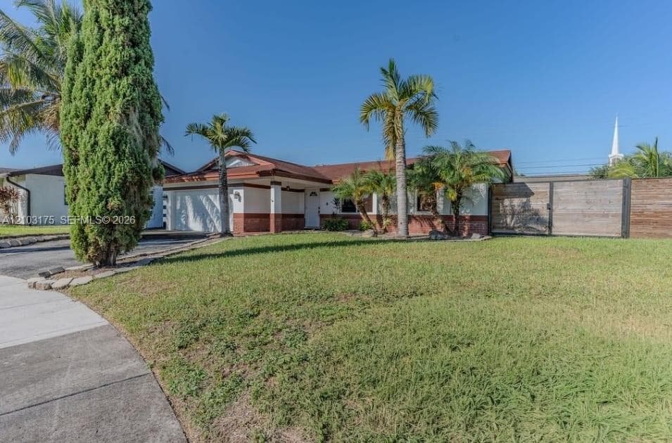 Single-story exterior featuring a brick facade, palm tree landscaping, a paved driveway, and horizontal wood-panel fencing