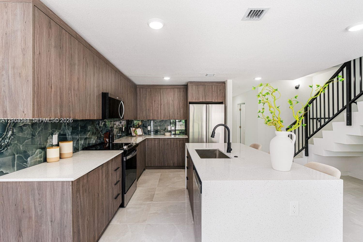 Kitchen featuring wood-finish cabinetry, white quartz countertops, stone-patterned backsplash, stainless steel appliances, and a central island with a matte black faucet