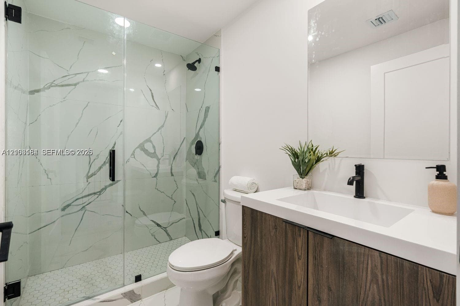 Bathroom featuring a frameless glass shower with floor-to-ceiling stone-patterned tile, a wood-finish vanity with an integrated countertop sink, matte black fixtures, and a large wall-mounted mirror